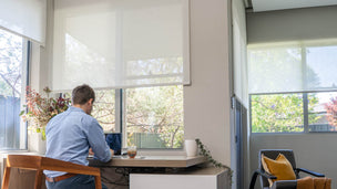 Person working at a desk with large windows and blinds in a bright room.