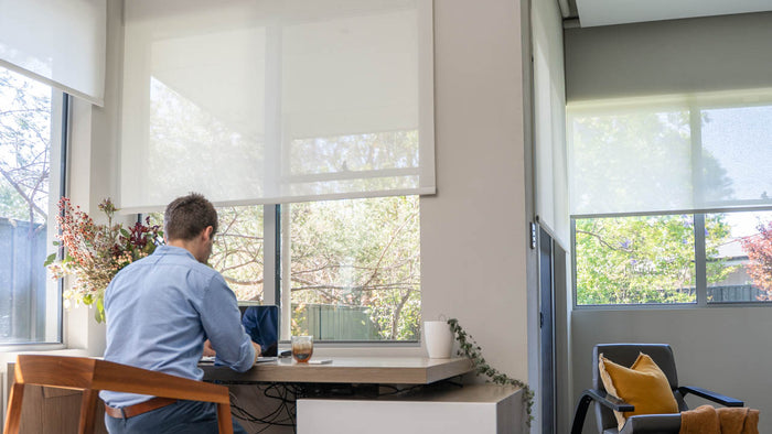 Person working at a desk with large windows and blinds in a bright room.